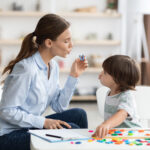 Young woman speech therapist teaching little boy with pronounciation deffects to say sound U during personal training at classroom