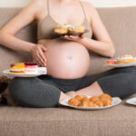 Close up of hungry pregnant woman sitting on the sofa is eating a lot of unhealthy food such as donuts, cakes, chocolate cereal balls, croissants and cookies. Sweet cravings during pregnancy concept.