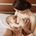 Young beautiful naked mom breastfeeding hugging her newborn baby smiling sitting on bed at home. Copy space.