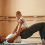 Happy athletic mother having fun with her baby while doing post-pregnancy exercises on the floor.