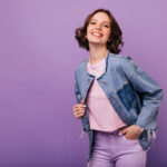 Studio portrait of confident young woman in trendy oversize jacket. Elegant smiling girl posing with pleasure on violet background.