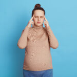 Young beautiful pregnant woman with closed eyes expecting baby, keeping fingers ob temples, thinks about something important, poses isolated on blue background, being confused and annoyed.