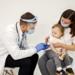 Male doctor in white medical gown is going to do vaccination. Boy and woman wearing white t-shirts. Doctor and mother wearing face masks.
