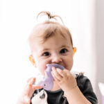 Cheerful baby girl with dark eyes looking at camera and sucking rubber toy while playing in light playroom at home