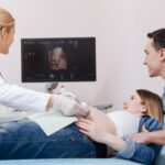 Admiring first digital image of our child. Delighted attentive professional obstetrician working in the sonography cabinet while examining pregnant belly and using ultrasound linear probe