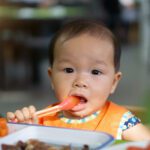 Asian baby girl in restaurant with a spoon eats food.