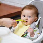 A nine month old smiling baby sits at a white table in a highchair and eats with a spoon from a bowl. Mom feeds the baby from a spoon. Blurred background. Healthy food for children. Children food.