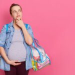 Pregnant woman thinking while standing isolated over pink background, looking away and keeps palm on chin, wearing casual attire, touching her belly, holding two bags with stuff, copy space for ad.