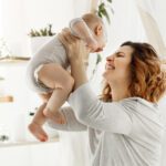 Happy smiling mother playing with newborn child in comfy light bedroom in front of window. Moments of motherhood happiness with kids. Family concept