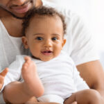 Portrait Of Adorable Black Baby Boy Spending Time With Father At Home, Happy Little Infant Child Relaxing In Daddy's Arms And Looking At Camera With Curiosity, Closeup Shot With Free Space, Cropped