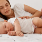 Young adult female with dark hair lying with baby in bed, looking at daughter to sees sleeping she or not, woman wearing white casual t shirt, happy motherhood.