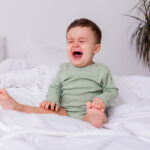 crying baby in a green cotton bodysuit and bare feet is sitting on a bed spread out. High quality photo